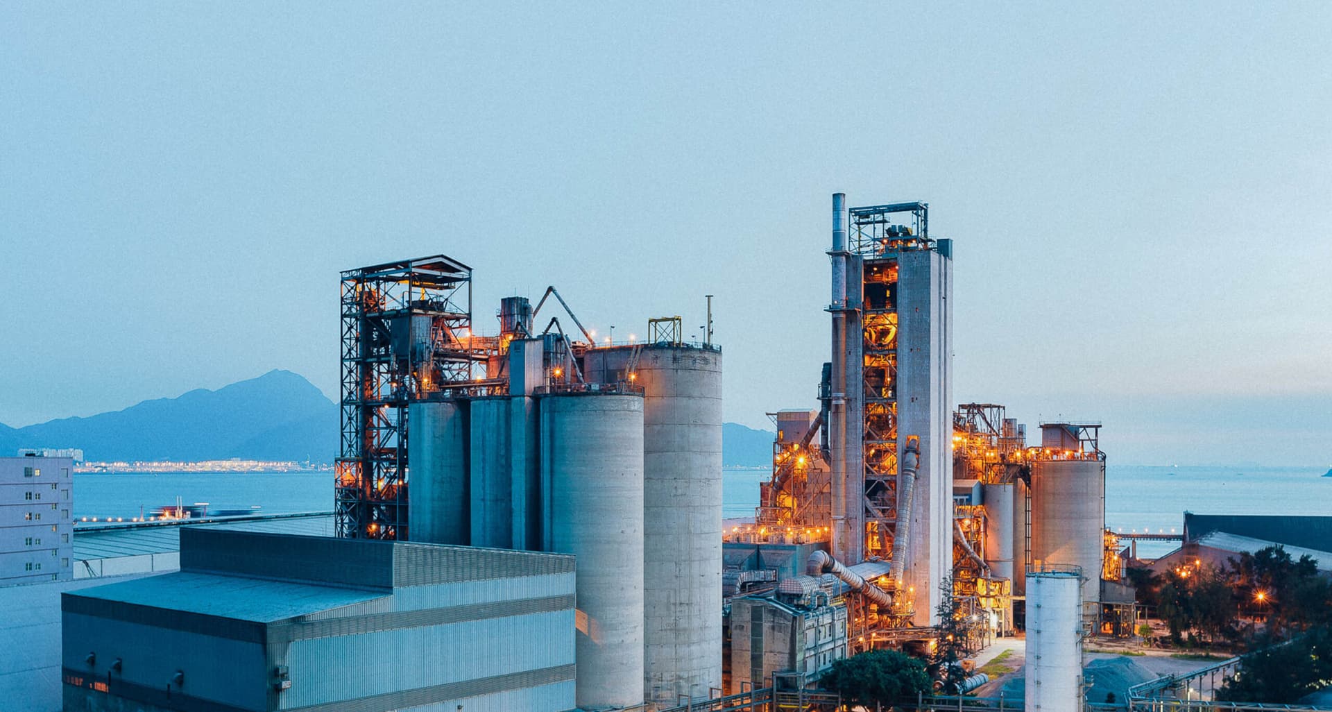 Industrial facility at dusk with illuminated silos and processing towers near a waterfront, mountains visible in the background. for Bo Kemp podcast.